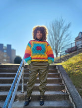 Load image into Gallery viewer, Black woman with her hair in a halo from the sun wearing a crocheted rainbow sweater and olive green fake leather pants.