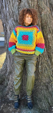 Load image into Gallery viewer, Black woman standing next to a large tree wearing a crocheted rainbow sweater and olive green fake leather pants.