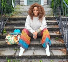 Load image into Gallery viewer, Black woman with a turquoise hat on top of a Southwestern style back. She is wearing a white sweater and crochet pants in grey, green, red, orange, gold and turquoise.