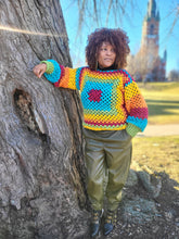 Load image into Gallery viewer, Black woman standing next to a large tree wearing a crocheted rainbow sweater and olive green fake leather pants. 
