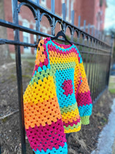 Load image into Gallery viewer, Colorful rainbow sweater hanging from a black velvet hanger with a gold hook hanging on a black wrought iron fence in front of a church 
