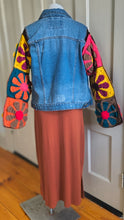 Load image into Gallery viewer, A denim jacket with colorful crocheted patchwork sleeves, shown from the back on a dress form with a rust-colored skirt. The jacket features bright pink, yellow, orange, and blue crochet patterns on the sleeves.
