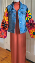 Load image into Gallery viewer, A cropped blue denim jacket with colorful crocheted flower patchwork sleeves, styled over a rust-orange turtleneck dress on a mannequin in an indoor setting.
