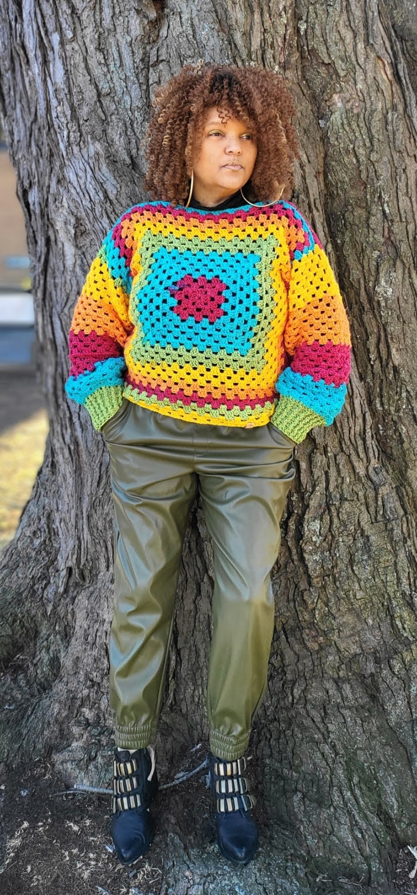 Black woman standing next to a large tree wearing a crocheted rainbow sweater and olive green fake leather pants. 