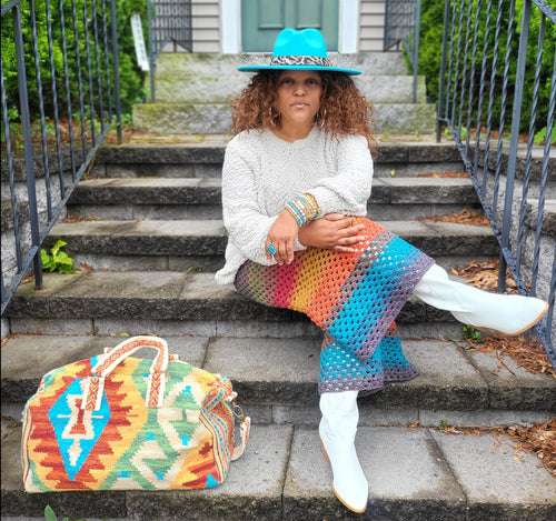 Black woman wearing a turquoise hat with a white sweater, a Southwestern bag and crochet pants in grey, green, red, orange, gold and turquoise. 