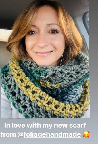 Woman wearing a hand crocheted scarf that is green grey and gold in color. The woman had light brown, blond hair and brown eyes. 